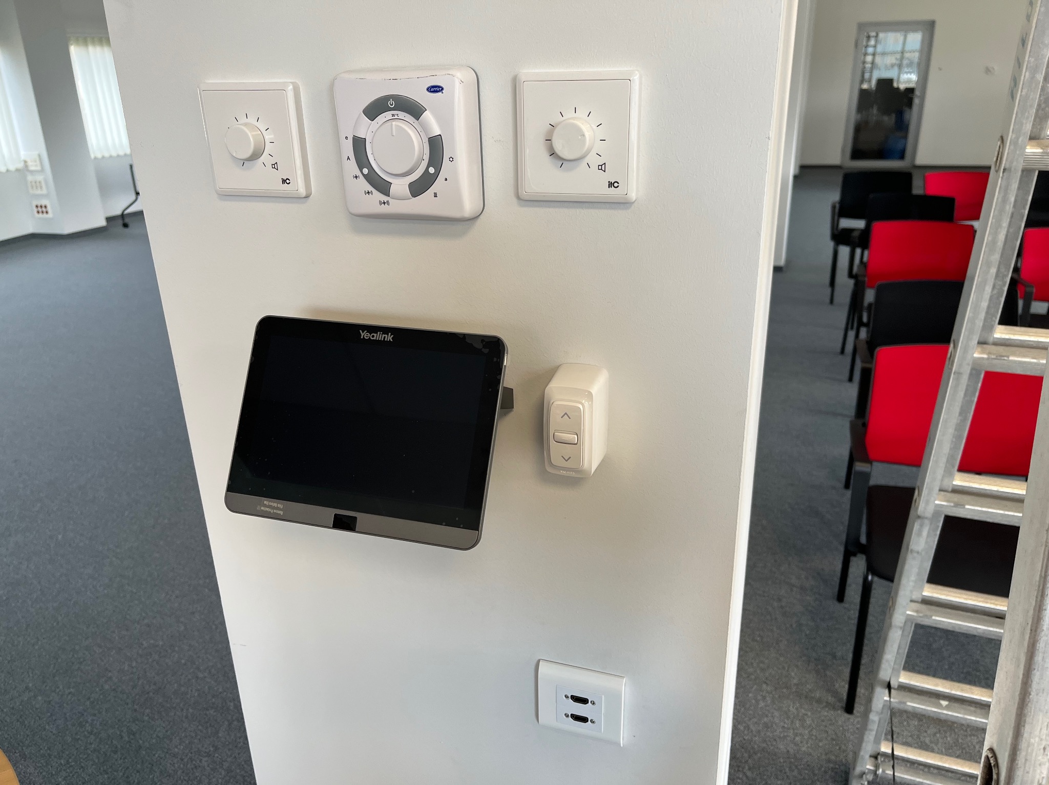 Panel sterowania Yealink z dotykowym ekranem, regulatorami głośności i gniazdami HDMI zamontowany na białej ścianie w sali konferencyjnej z widocznymi krzesłami i drabiną.