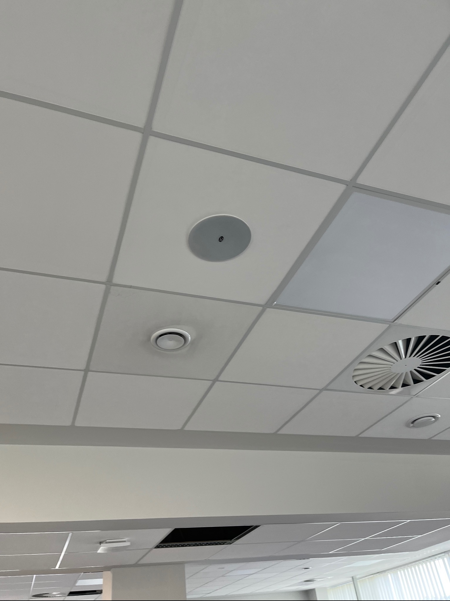 Kasetonowy sufit podwieszany z okrągłym głośnikiem sufitowym, okrągłym czujnikiem i kratką wentylacyjną.
