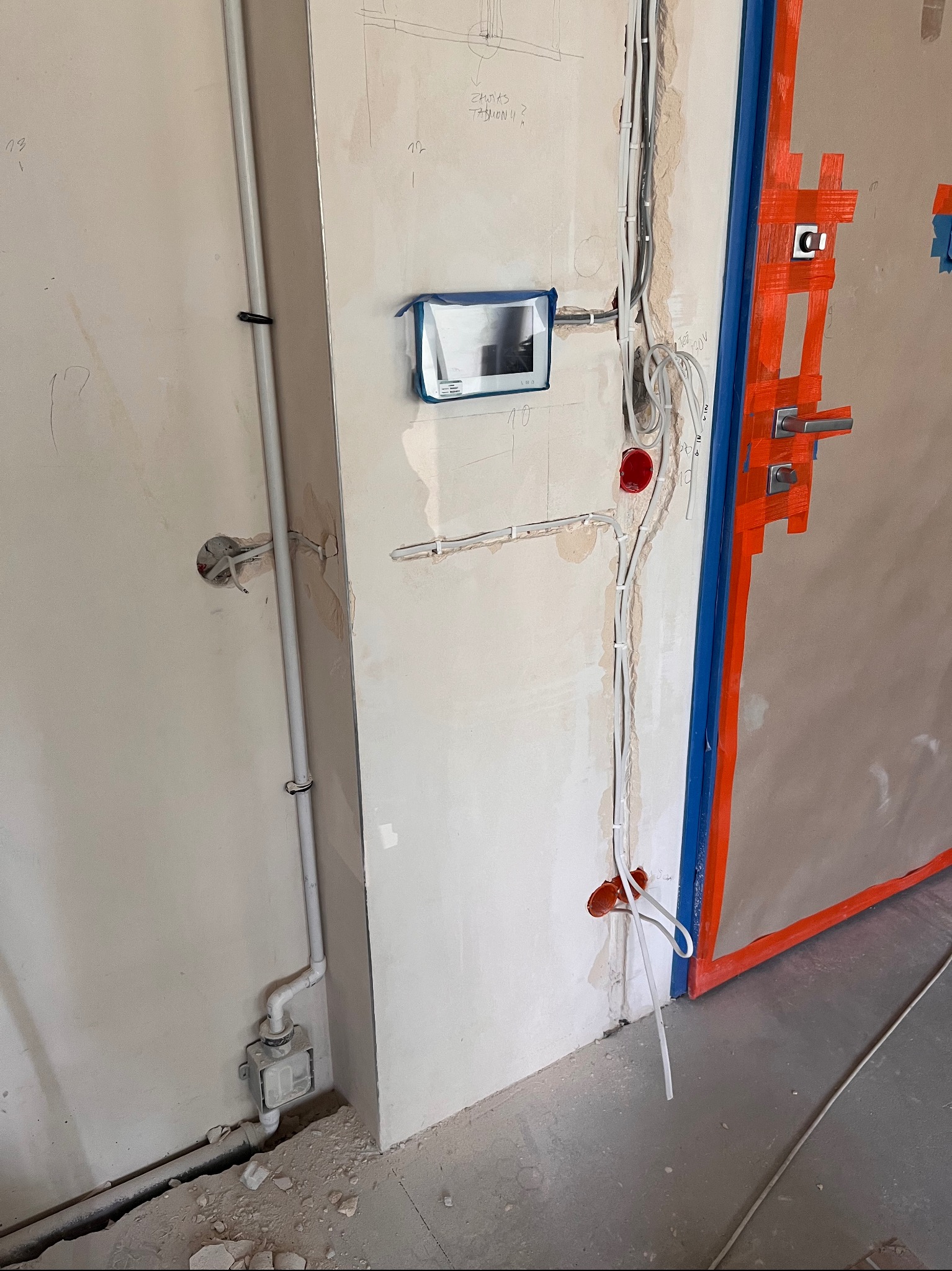 Instalacja elektryczna w trakcie montażu: przewody, puszki i elementy domofonu na ścianie w surowym stanie, zabezpieczone drzwi w tle. Widoczne szkice instalacji na ścianie.