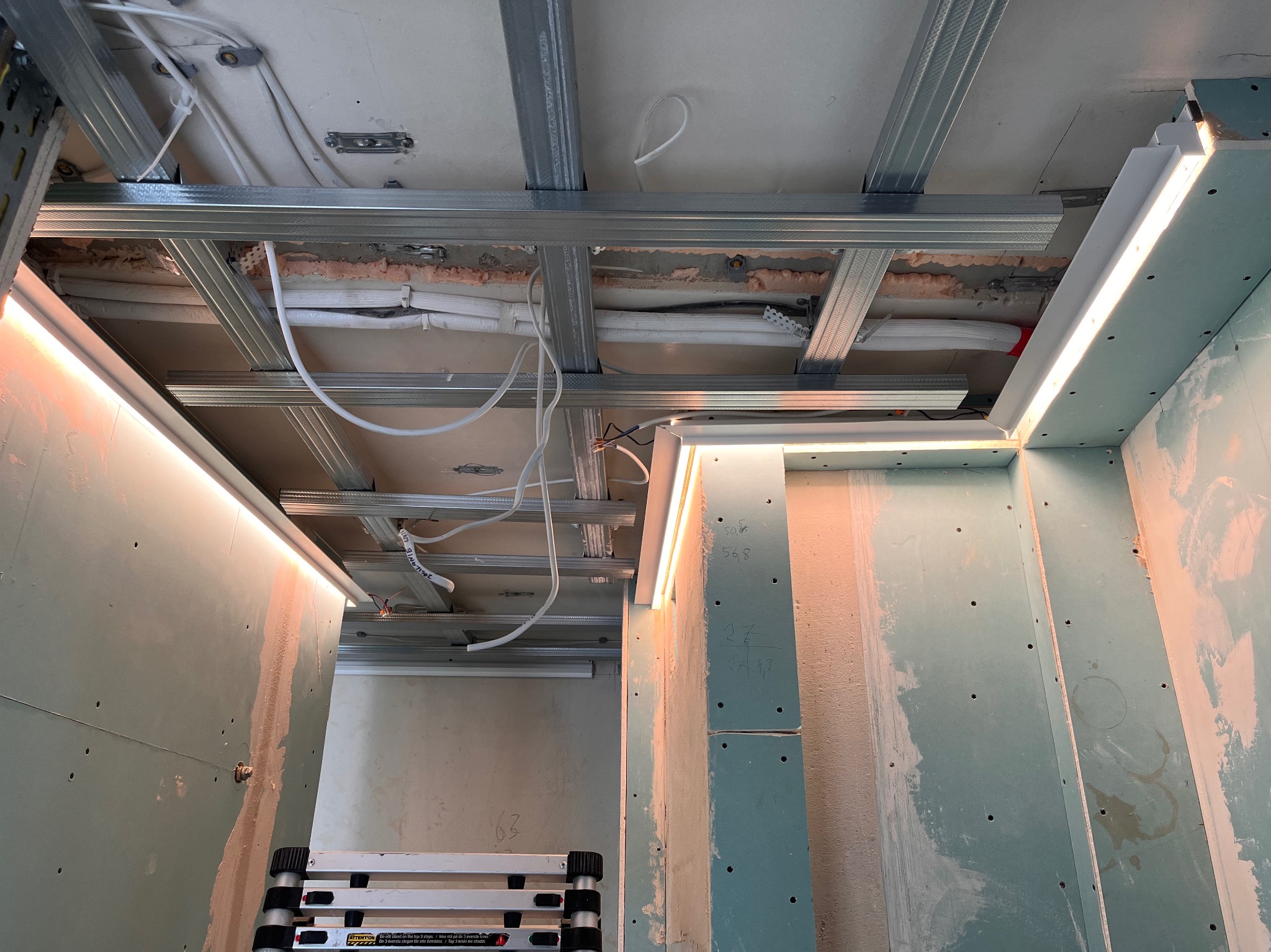 Montaż sufitu podwieszanego z oświetleniem LED w pomieszczeniu. Widoczne kable, profile i płyty gipsowo-kartonowe. Praca w toku, drabina w kadrze.