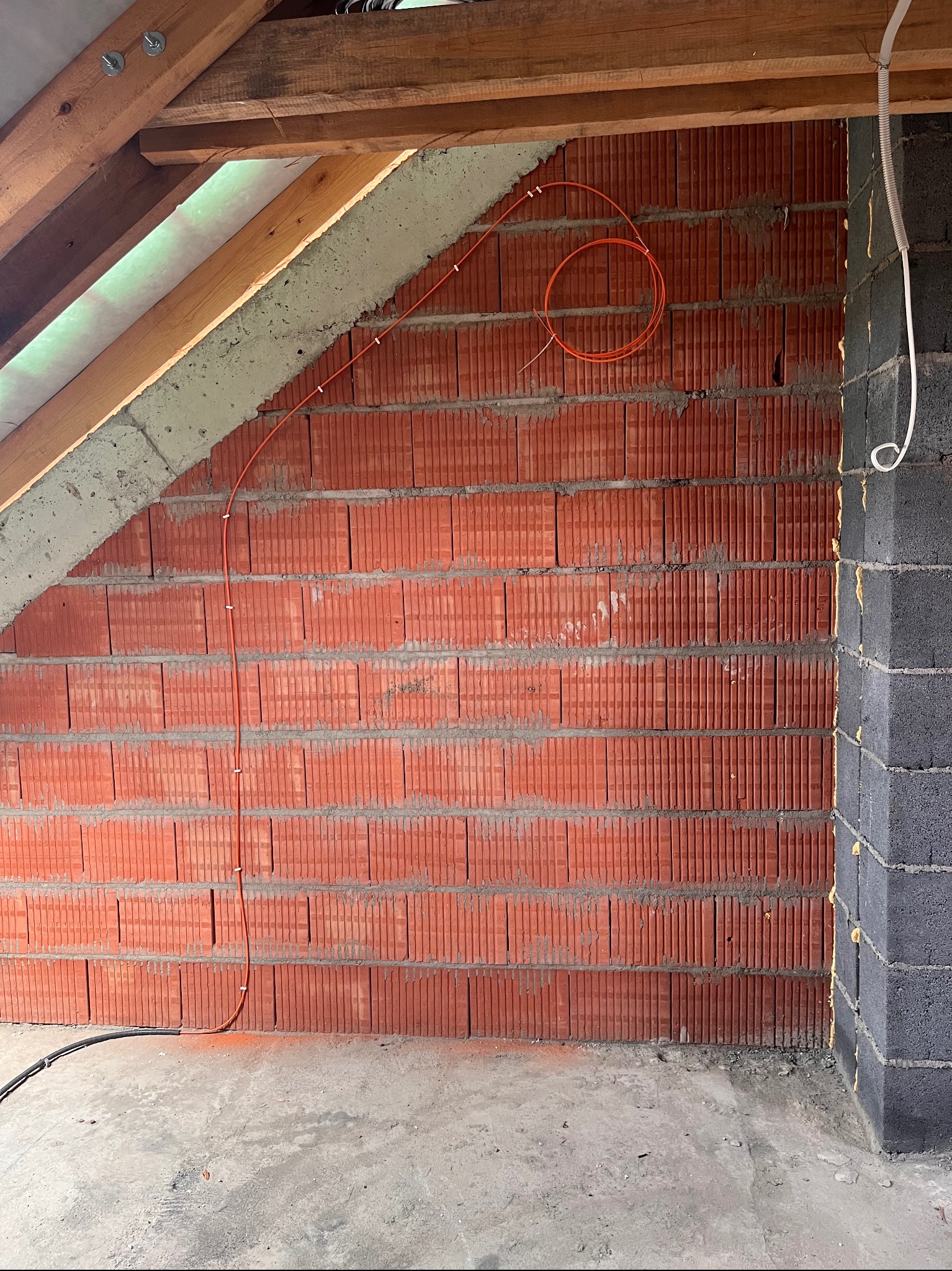Czerwona cegła z pomarańczowym przewodem w kształcie pętli, przymocowanym klamrami. Fragment poddasza z drewnianą konstrukcją w tle. Szara ściana z pustaków po prawej.