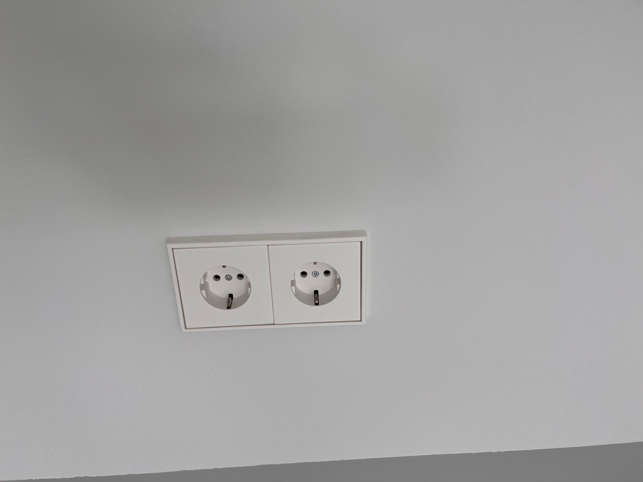 Białe, podwójne gniazdko elektryczne z uziemieniem, zamontowane na jasnej ścianie. Widok z dołu, minimalizm i symetria w ujęciu.