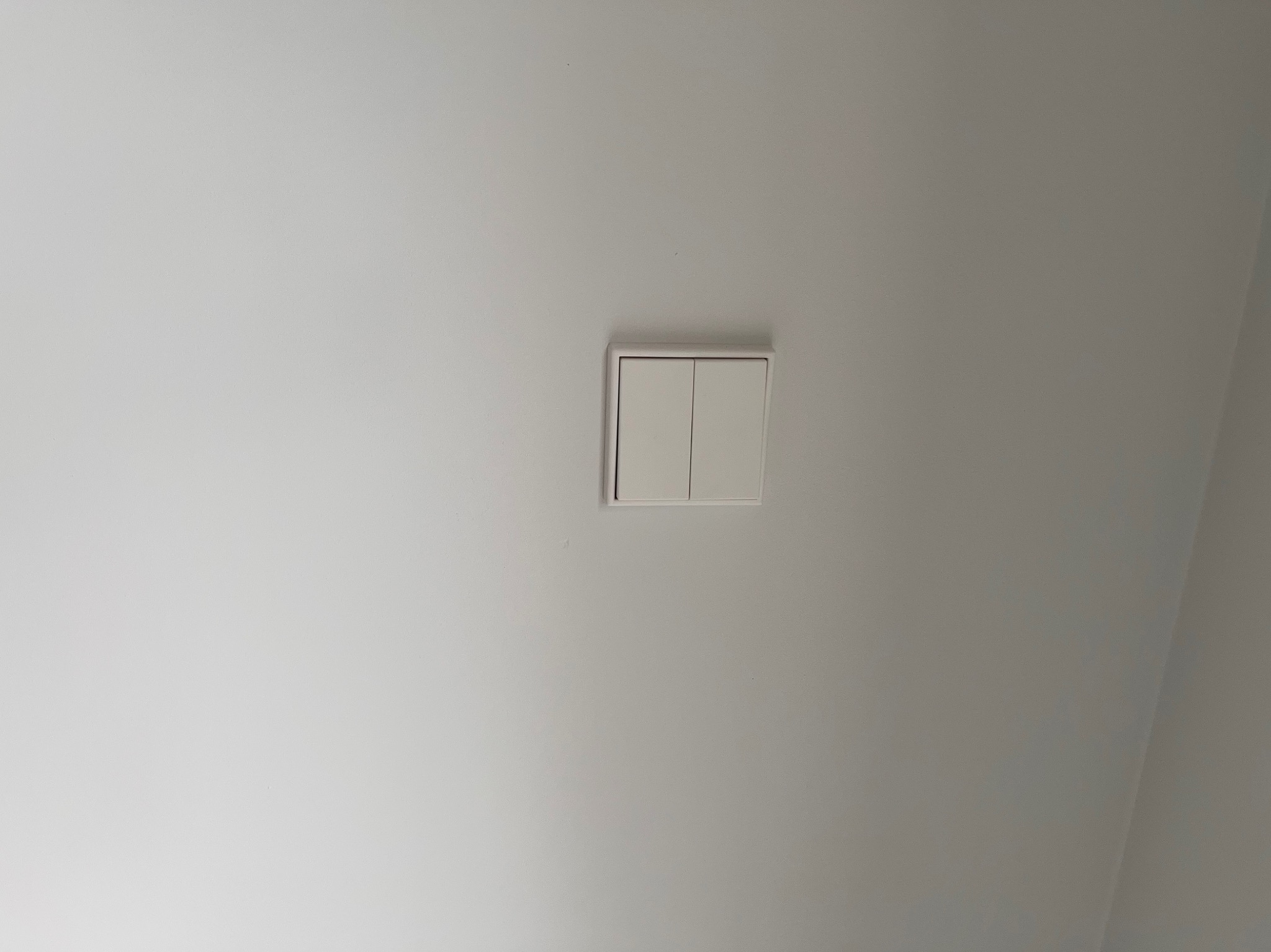 Biały, podwójny włącznik światła na gładkiej, białej ścianie, zamontowany tuż pod sufitem. Minimalistyczny design w nowoczesnym wnętrzu.