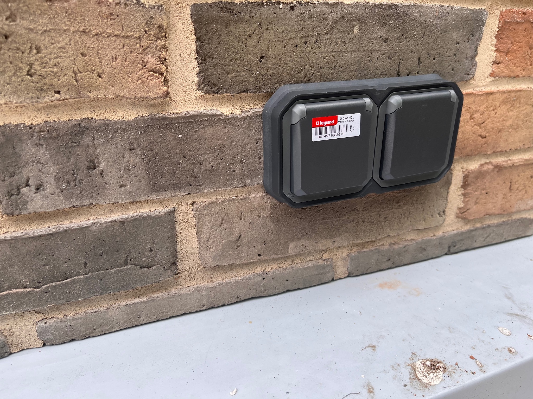 Zewnętrzne, podwójne gniazdko elektryczne Legrand, zamontowane na ceglanej ścianie. Widoczna etykieta producenta na górnej klapce.
