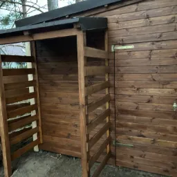 Brązowa, drewniana konstrukcja z zadaszeniem, prawdopodobnie składzik na drewno, w trakcie budowy, widoczny pracownik w odzieży roboczej i narzędzia.