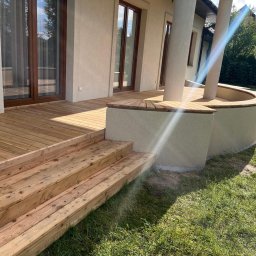 DREWBET SP. Z O.O. - Drewniany taras przy domu z drewnianymi schodami i półokrągłymi siedziskami z drewna, wspartymi na kolumnach. Widok na trawnik.