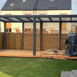 DREWBET SP. Z O.O. - Nowoczesna pergola z drewnianym tarasem i ażurowymi ścianami, w tle budynek mieszkalny. Elementy konstrukcyjne w kolorze grafitowym. Widoczne narzędzia i materiały budowlane.