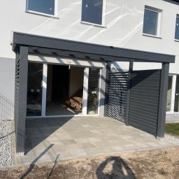 DREWBET SP. Z O.O. - Nowoczesna, szara pergola z lamelami i wyłożony kostką taras przylegający do budynku. Widoczne okna i ogrodzenie z siatki.