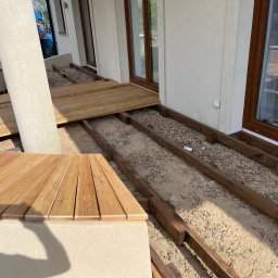 DREWBET SP. Z O.O. - Montaż drewnianego tarasu: widoczne legary, częściowo ułożone deski i zabudowa siedziska. Etap budowy tarasu przy budynku z oknami balkonowymi.