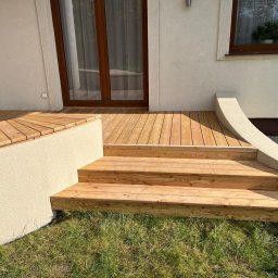 DREWBET SP. Z O.O. - Nowy taras z drewna z niskimi schodami wejściowymi, obok domu z oknem i drzwiami balkonowymi, otoczony trawnikiem. Jasne drewno i elewacja.