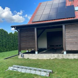 DREWBET SP. Z O.O. - Drewniana pergola z żaluzjami w trakcie budowy, widoczne panele słoneczne na dachu domu i drabina na trawniku. Ujęcie z niskiego kąta.