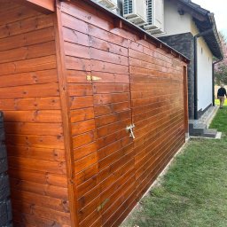 DREWBET SP. Z O.O. - Lakierowana na brązowo drewniana konstrukcja z drzwiami na posesji, obok czarne zbiorniki na deszczówkę. W tle dom z klimatyzacją i osoba na trawniku.