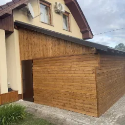 Drewniana zabudowa przylegająca do budynku z ciemnymi drzwiami i zadaszeniem, komponująca się z elewacją domu i otoczeniem. Widok z boku, z elementami zieleni w kadrze.