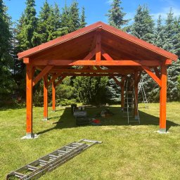 DREWBET SP. Z O.O. - Drewniana altana w trakcie budowy, widoczne narzędzia i drabina, zielone otoczenie z drzewami iglastymi, słoneczny dzień. Elementy konstrukcyjne w kolorze pomarańczowym.