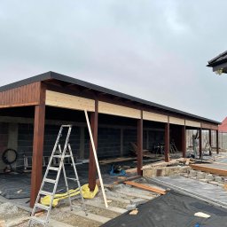 DREWBET SP. Z O.O. - Budowa zadaszenia w Marcinkowicach: drewniane słupy, czarny dach, widoczna drabina i narzędzia. Jeden pracownik przy pracy, drugi w tle. Etap konstrukcyjny.