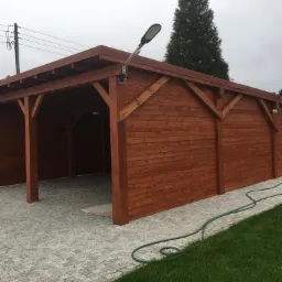 Drewniana altana z oświetleniem zewnętrznym i zielonym wężem ogrodowym na brukowanym podłożu. Konstrukcja pomalowana na ciepły, brązowy kolor, widok z boku.