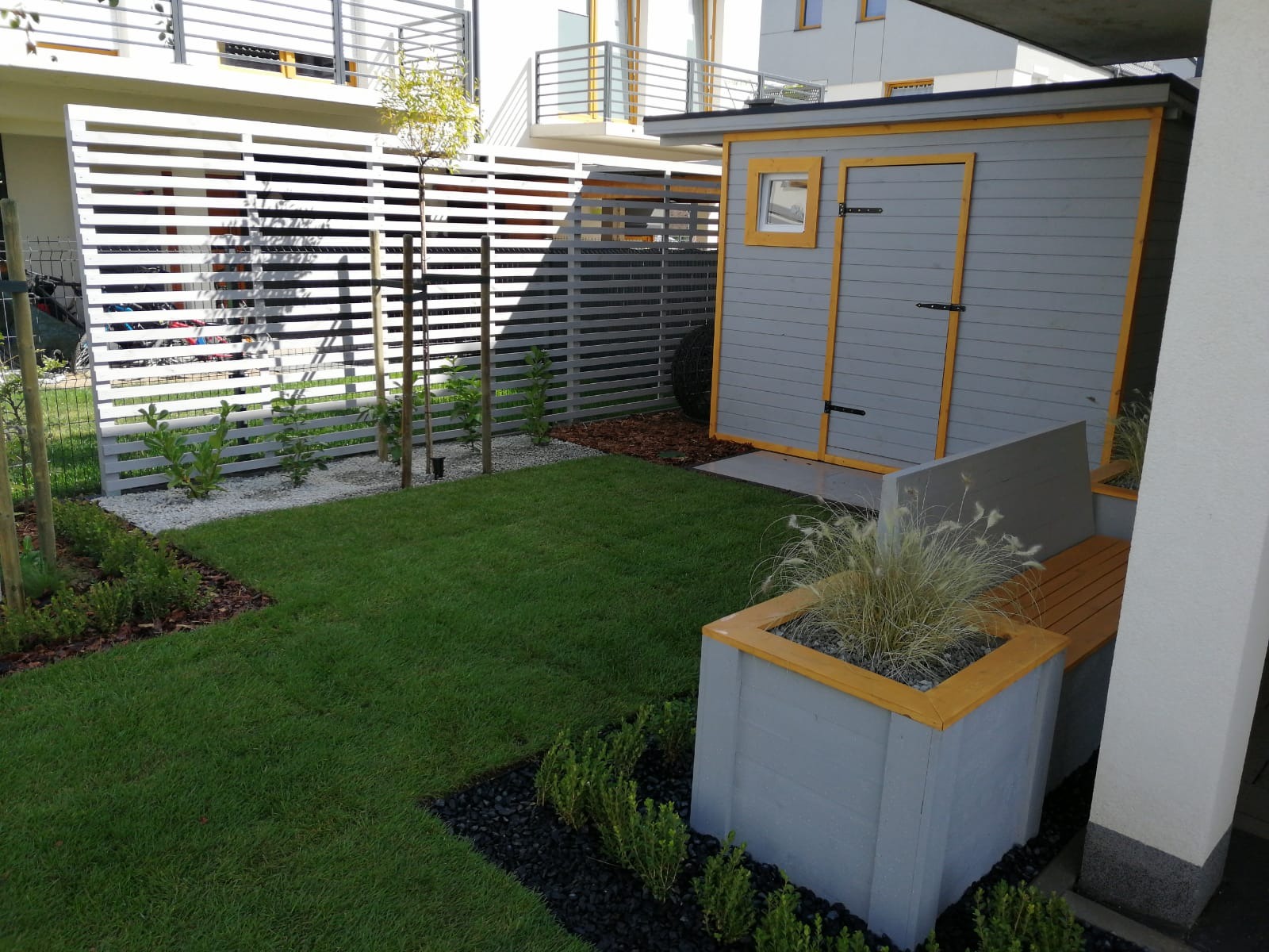 Nowoczesna aranżacja ogrodu z szarą altaną z żółtymi obramowaniami, drewnianą ławeczką w donicy i ażurowym, białym ogrodzeniem. Zadbane trawniki i roślinność.