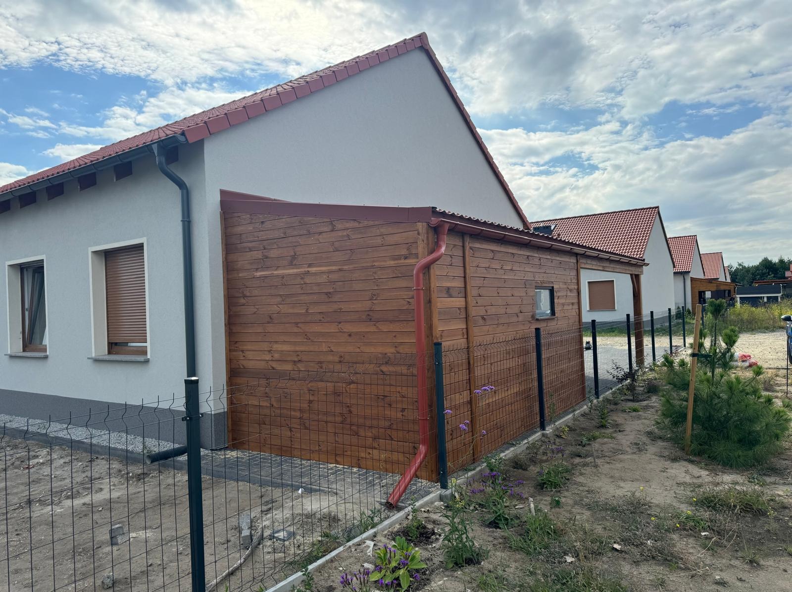 Drewniana zabudowa ściany domu z brązowymi rynnami, widok z zewnątrz. Ogrodzenie z siatki, trawa i inne domy w tle.