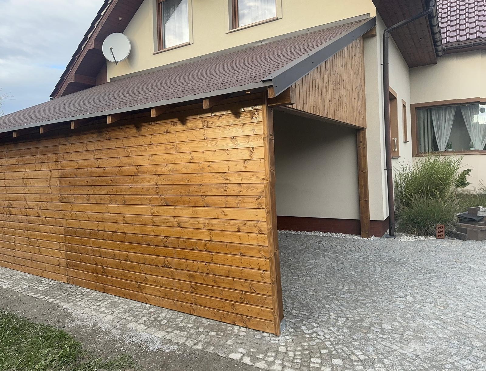 Drewniana konstrukcja z brązowym dachem, przylegająca do budynku. Widoczna boczna ściana z desek i wjazd wyłożony kostką. Element zabudowy w przestrzeni przed domem.
