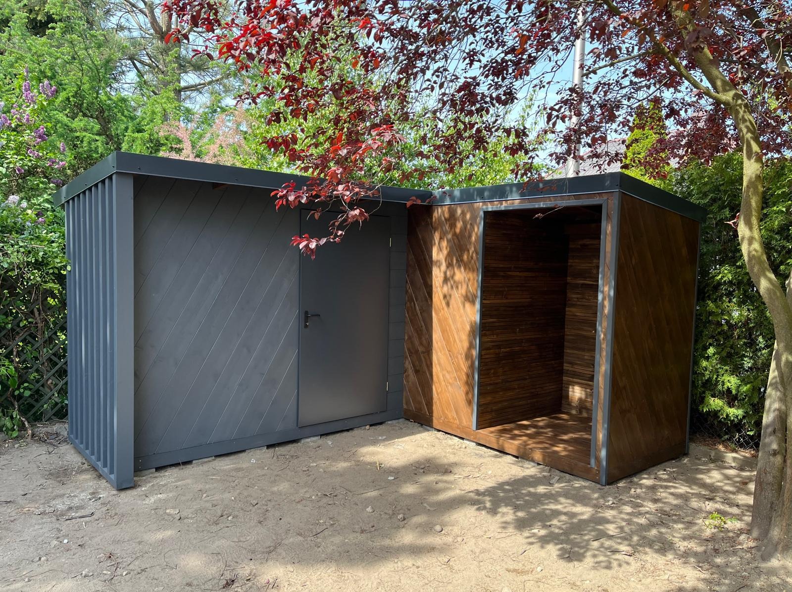 Nowoczesna, dwuczęściowa altana ogrodowa z drewna i metalu, z szarą, zamykaną częścią i otwartą, brązową przestrzenią, na tle zieleni i drzew z czerwonymi liśćmi.