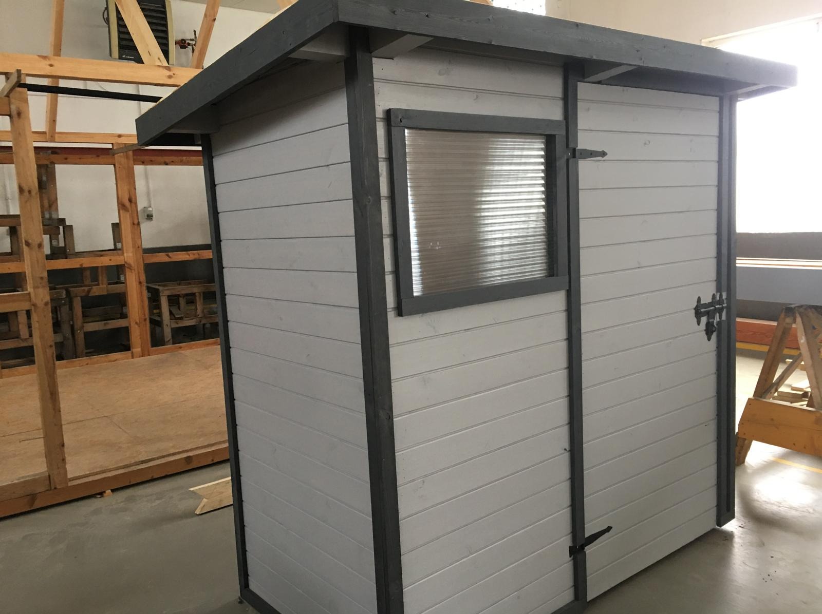 Szara, drewniana altana z oknem z tworzywa i ciemnymi detalami, w trakcie budowy, widoczny szkielet konstrukcji w tle. Prezentacja ukończonego projektu.