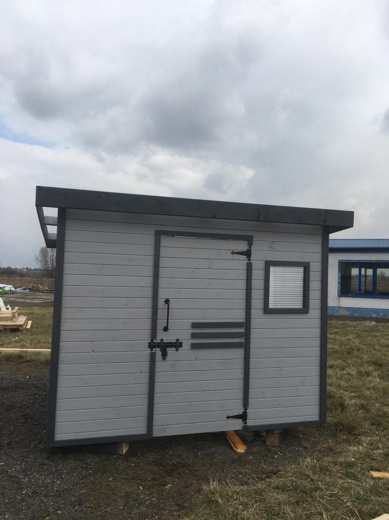 Szara, drewniana altana z drzwiami i małym oknem, umieszczona na trawiastym terenie pod pochmurnym niebem. Widoczne elementy konstrukcyjne i zamek.