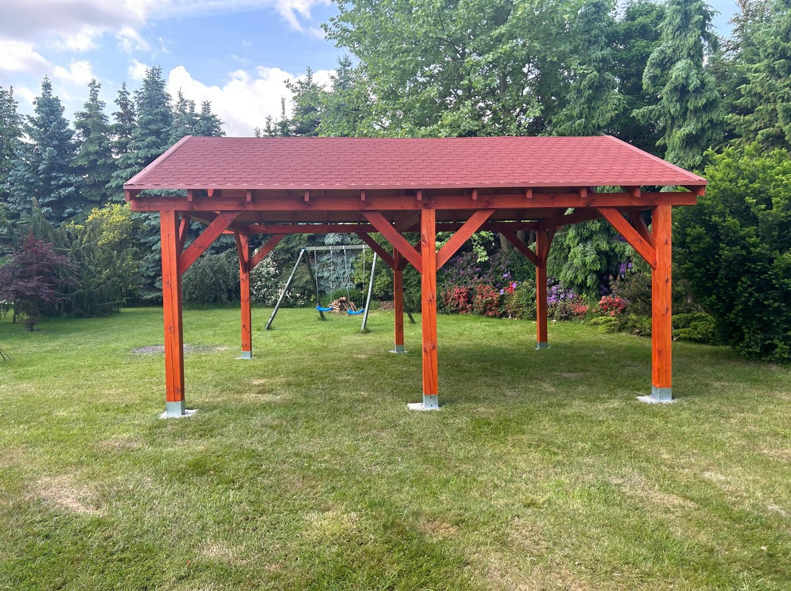 Nowoczesna, drewniana altana z czerwoną dachówką, osadzona na betonowych podstawach. W tle zielony trawnik, drzewa iglaste i huśtawka.