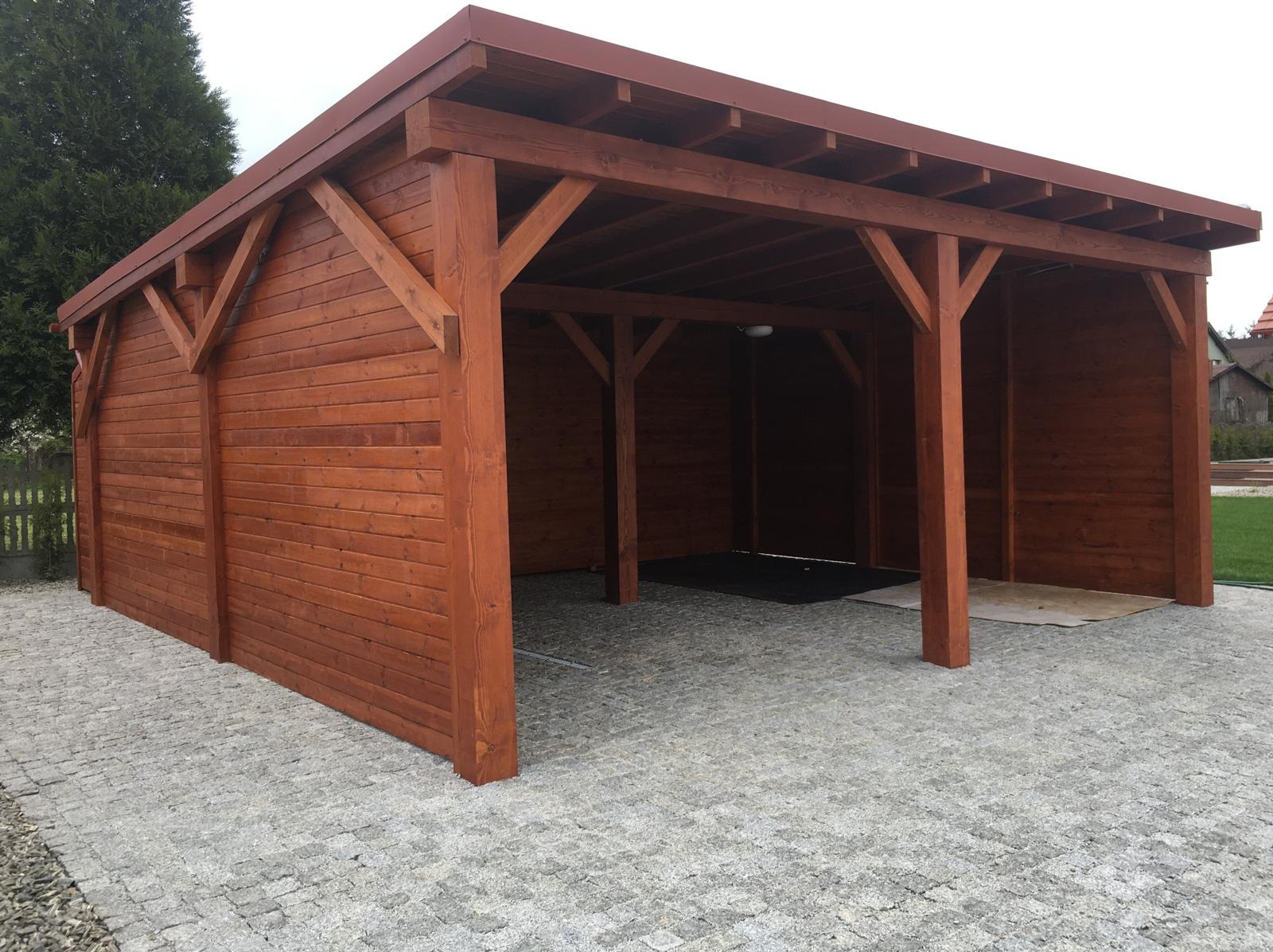 Drewniana altana zadaszona blachą, widok z zewnątrz. Konstrukcja z belek w kolorze brązowym, ściany z desek. Podłoże wyłożone kostką brukową.