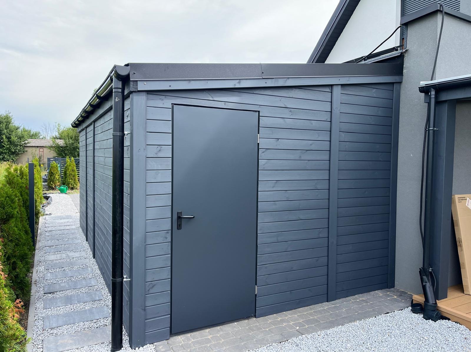 Nowoczesna, grafitowa altana z drewnianymi panelami i metalowymi elementami, umiejscowiona przy domu. Widoczny fragment ścieżki z kamieni.