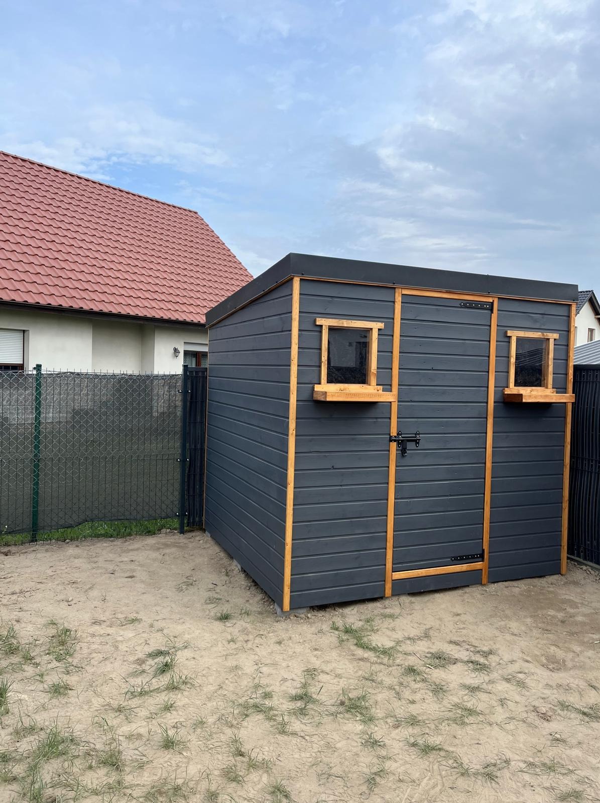 Nowoczesna, grafitowa altana ogrodowa z drewnianymi ramami okien i drzwi, widoczna na tle domu z czerwoną dachówką i zielonym ogrodzeniem.