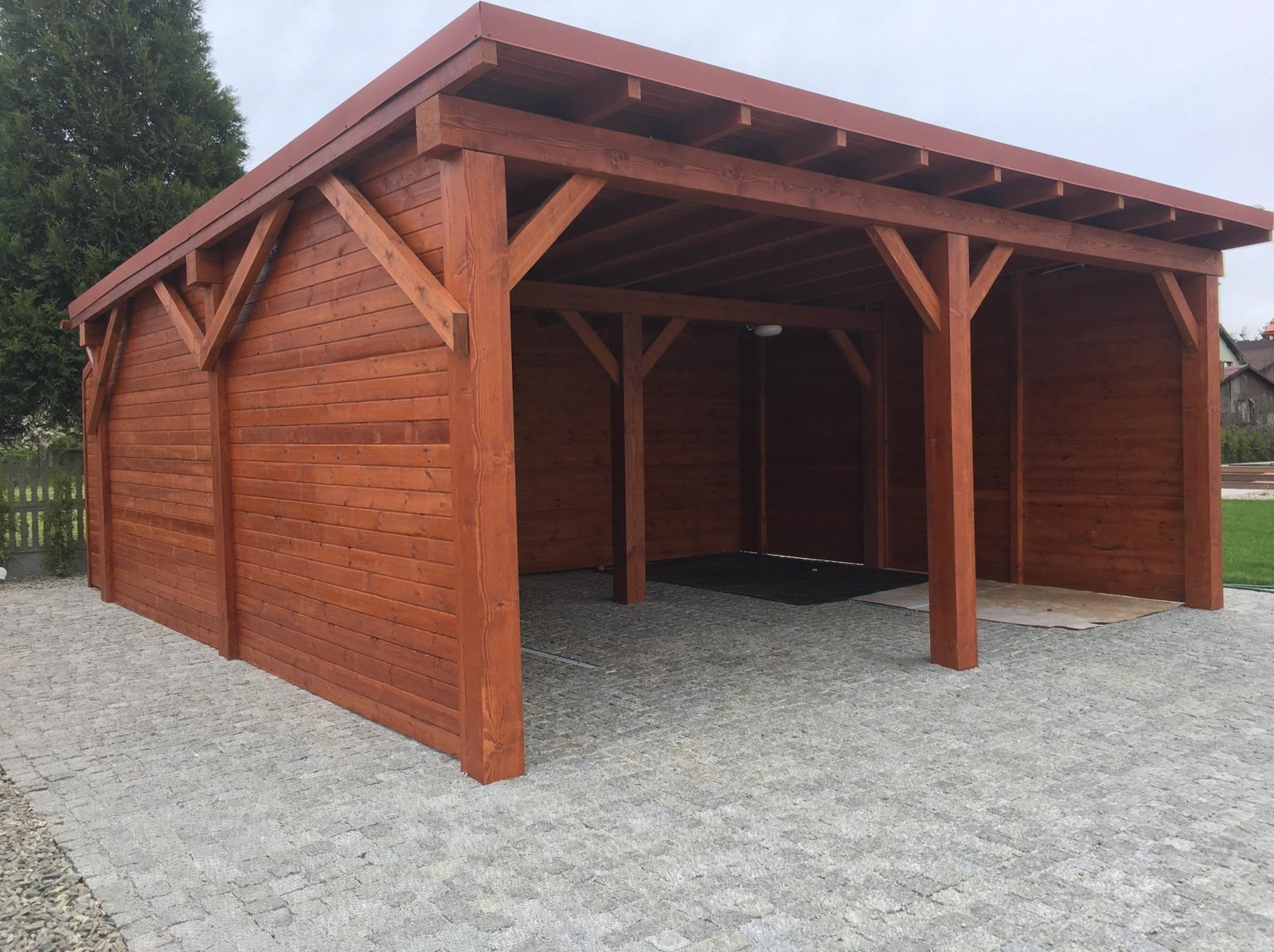 Drewniana wiata garażowa z brązowymi ścianami i dachem, na szarej kostce brukowej. Widoczne elementy konstrukcyjne i oświetlenie wewnątrz.