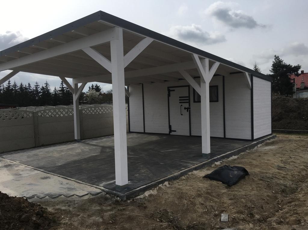 Biała drewniana pergola z szarą dachówką, przylegająca do budynku z szarej kostki brukowej. Widoczny betonowy płot i ziemia.