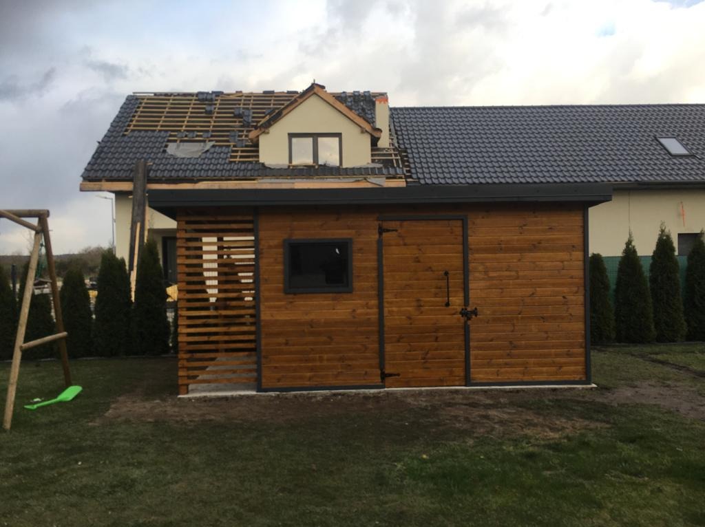 Drewniana altana z oknem i ażurową ścianą, na tle domu w trakcie remontu dachu, otoczona zielenią. Ujęcie z niskiej perspektywy, pochmurne niebo.