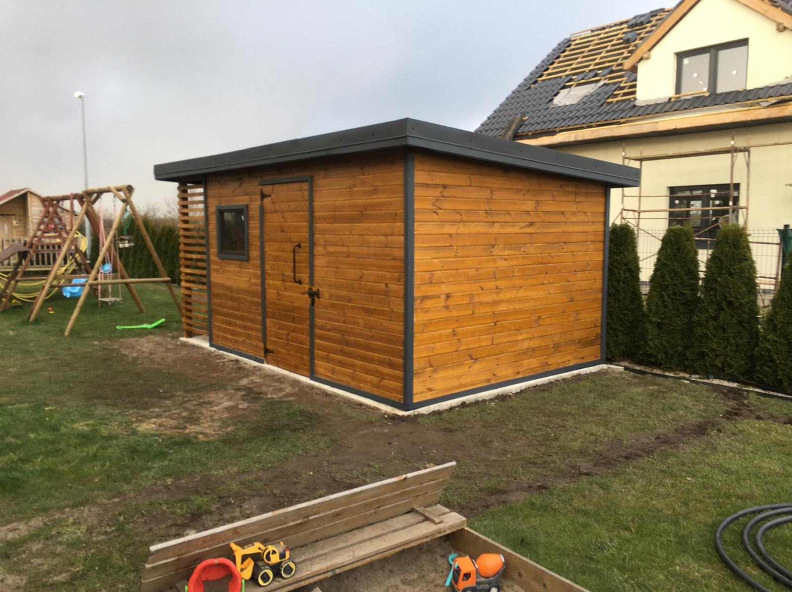 Drewniany domek narzędziowy z płaskim dachem i szarymi obramowaniami, na trawniku obok placu zabaw i domu w budowie. Widoczne zabawki w piaskownicy.