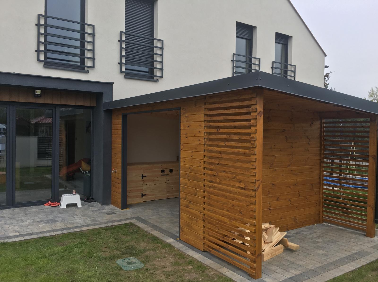Nowoczesna altana z drewna w kolorze miodowym, z ażurowymi ścianami, przylegająca do budynku z szarymi oknami i balkonami. Na ziemi kostka brukowa i trawa.