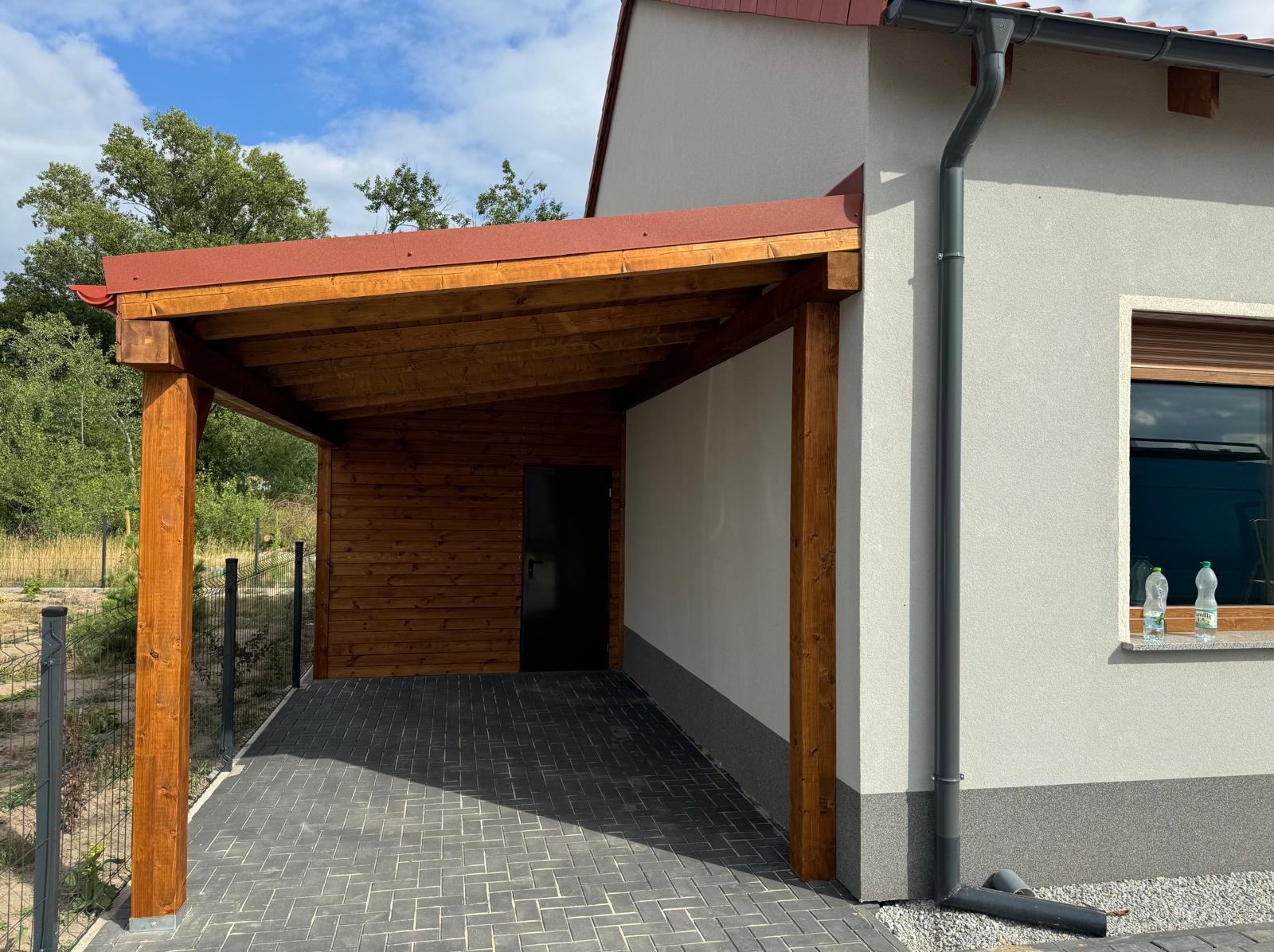 Drewniana wiata przy domu z czerwonym dachem, wsparta na solidnych słupach, z drewnianą zabudową ściany i brukowanym podjazdem. Widok na fragment budynku.