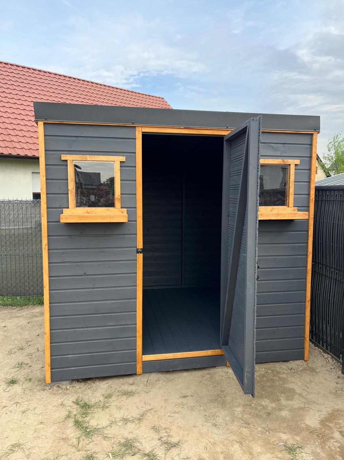 Nowoczesna, grafitowa altana ogrodowa z drewnianymi oknami i otwartymi drzwiami, na tle domu z czerwoną dachówką.