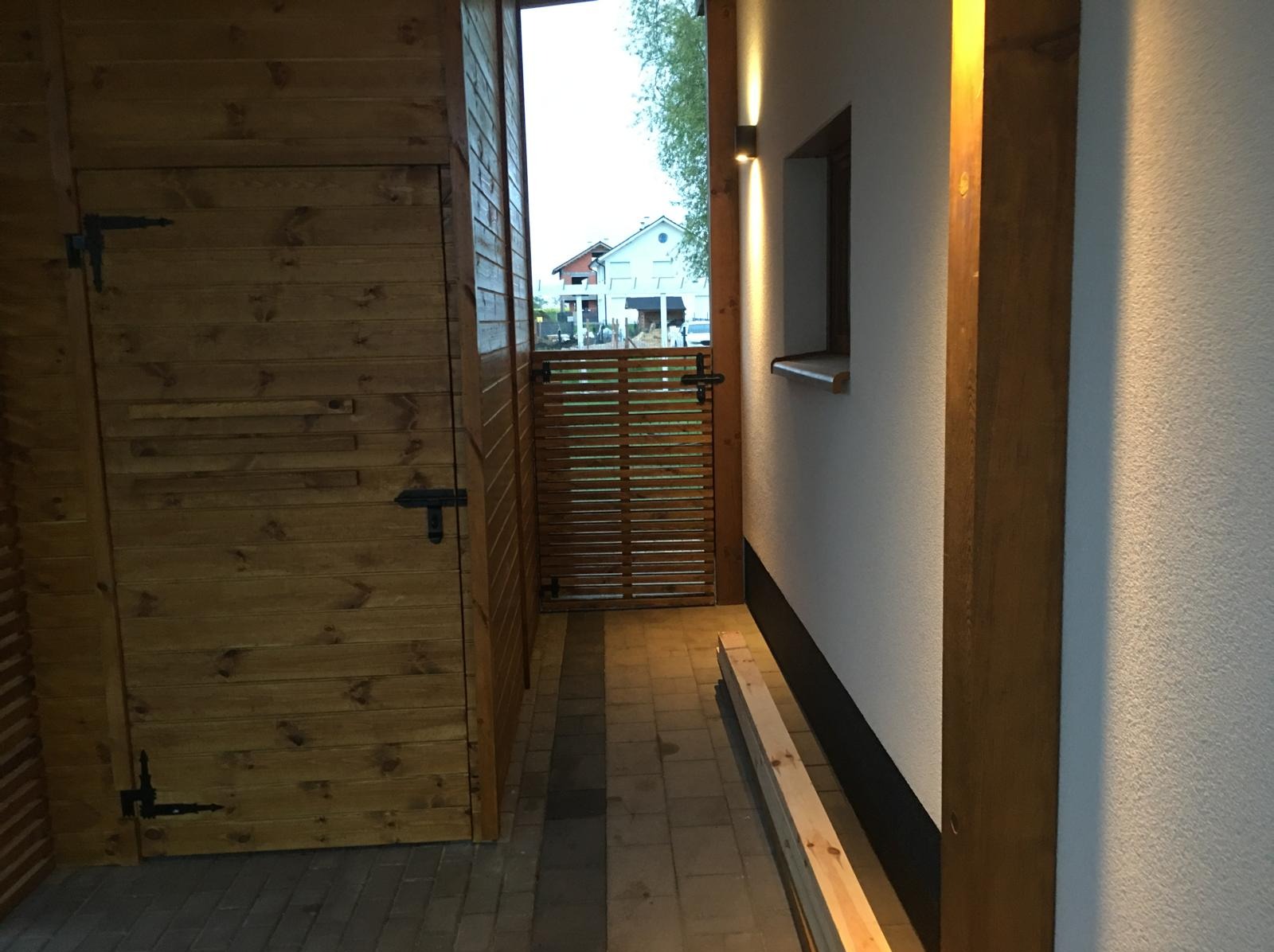 Drewniana zabudowa z furtką i oświetleniem zewnętrznym. Widok na dom przez furtkę. Elementy wykończeniowe z drewna, kostka brukowa.
