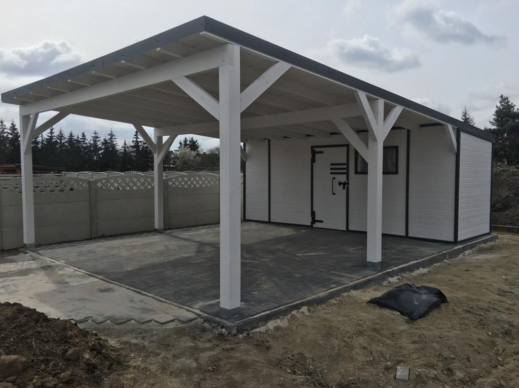 Biała drewniana altana z przylegającym budynkiem gospodarczym na kostce brukowej, otoczona ziemią i betonowym ogrodzeniem.