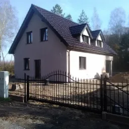 Dwukondygnacyjny dom z ciemnym, skośnym dachem i oknami, otoczony czarnym, kutym ogrodzeniem i drzewami w jesiennej scenerii.