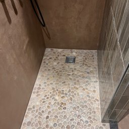 Domi-Bud - Kabina prysznicowa z podłogą wyłożoną drobnymi kamieniami w odcieniach beżu i brązu. Ściany wykończone w brązowym tynku i płytkach. Widoczny odpływ liniowy.