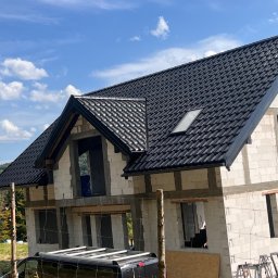 Domi-Bud - Dom w trakcie budowy z czarnym dachem z dachówki ceramicznej i białymi ścianami z betonu komórkowego, widok z dołu na tle błękitnego nieba.