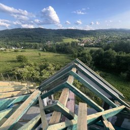 Domi-Bud - Konstrukcja dachu z drewnianych belek na tle malowniczego krajobrazu z zielonymi wzgórzami i błękitnym niebem z chmurami. Widok z góry na budowę.