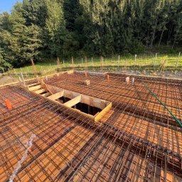 Domi-Bud - Zbrojenie fundamentów domu jednorodzinnego z drewnianym szalunkiem i wystającymi prętami. Widok z góry na przygotowany teren budowy w słoneczny dzień.