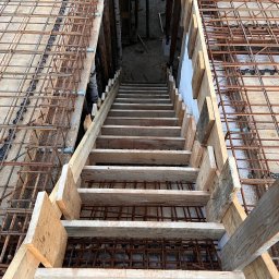 Domi-Bud - Zbrojenie stropu z drewnianym zejściem do piwnicy. Widok z góry na siatkę zbrojeniową i deskowanie. Etap prac budowlanych z użyciem stali i drewna.