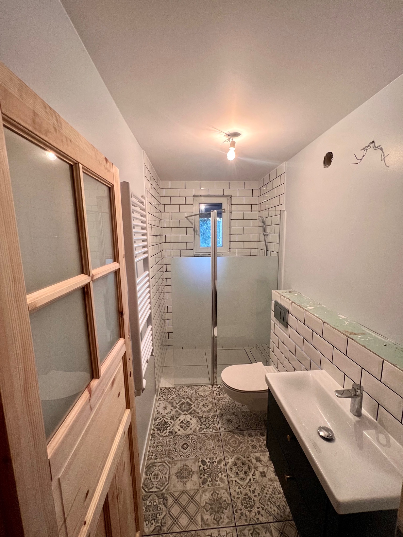 Nowoczesna łazienka w trakcie remontu z drewnianymi drzwiami z szybami, białą glazurą, wzorzystą podłogą i niedokończonymi elementami elektrycznymi.