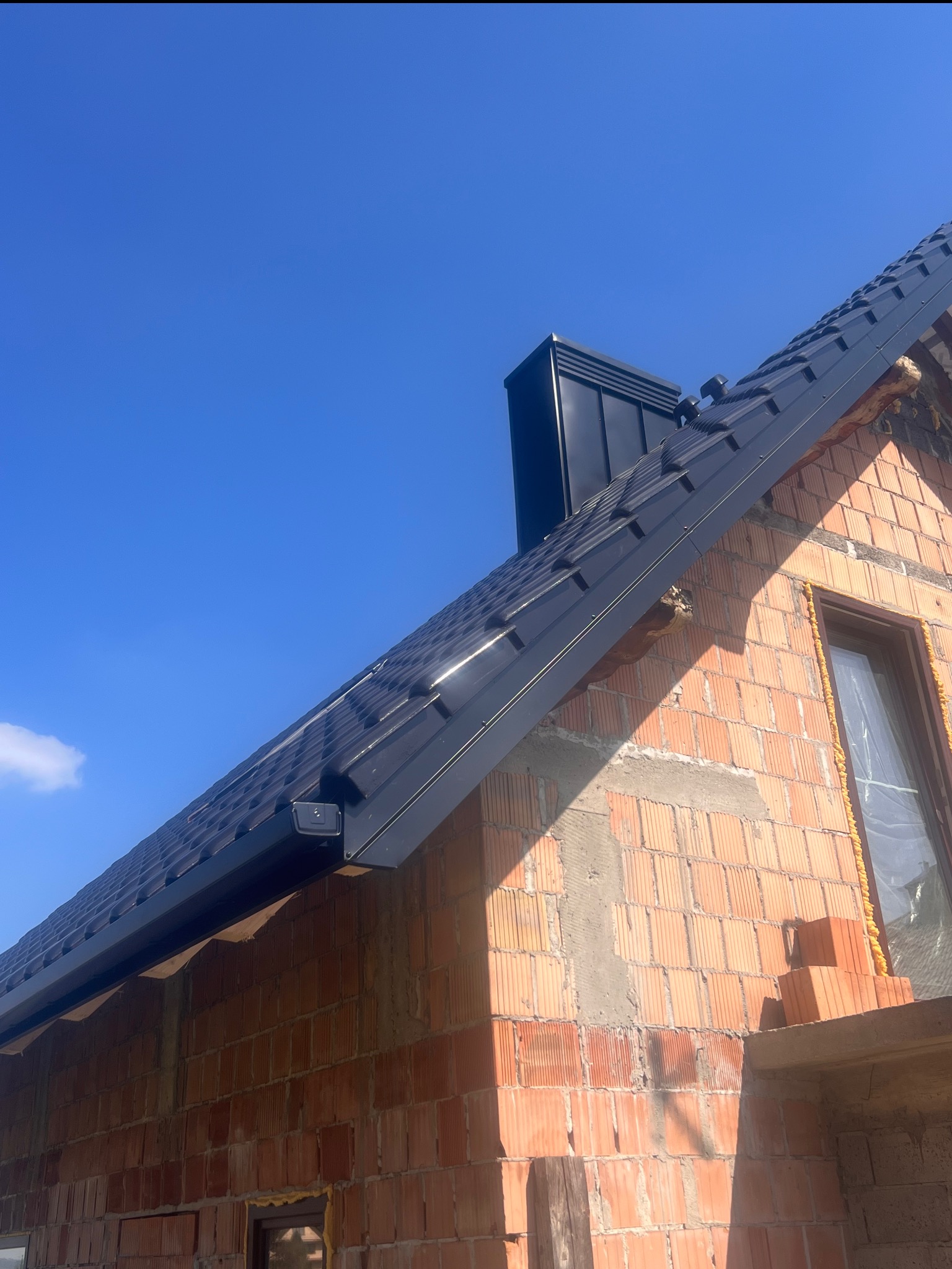 Surowa elewacja z cegły i czarny dach z blachy na tle błękitnego nieba. Widoczny komin i okno z pianką montażową. Ujęcie z dołu, w słoneczny dzień.