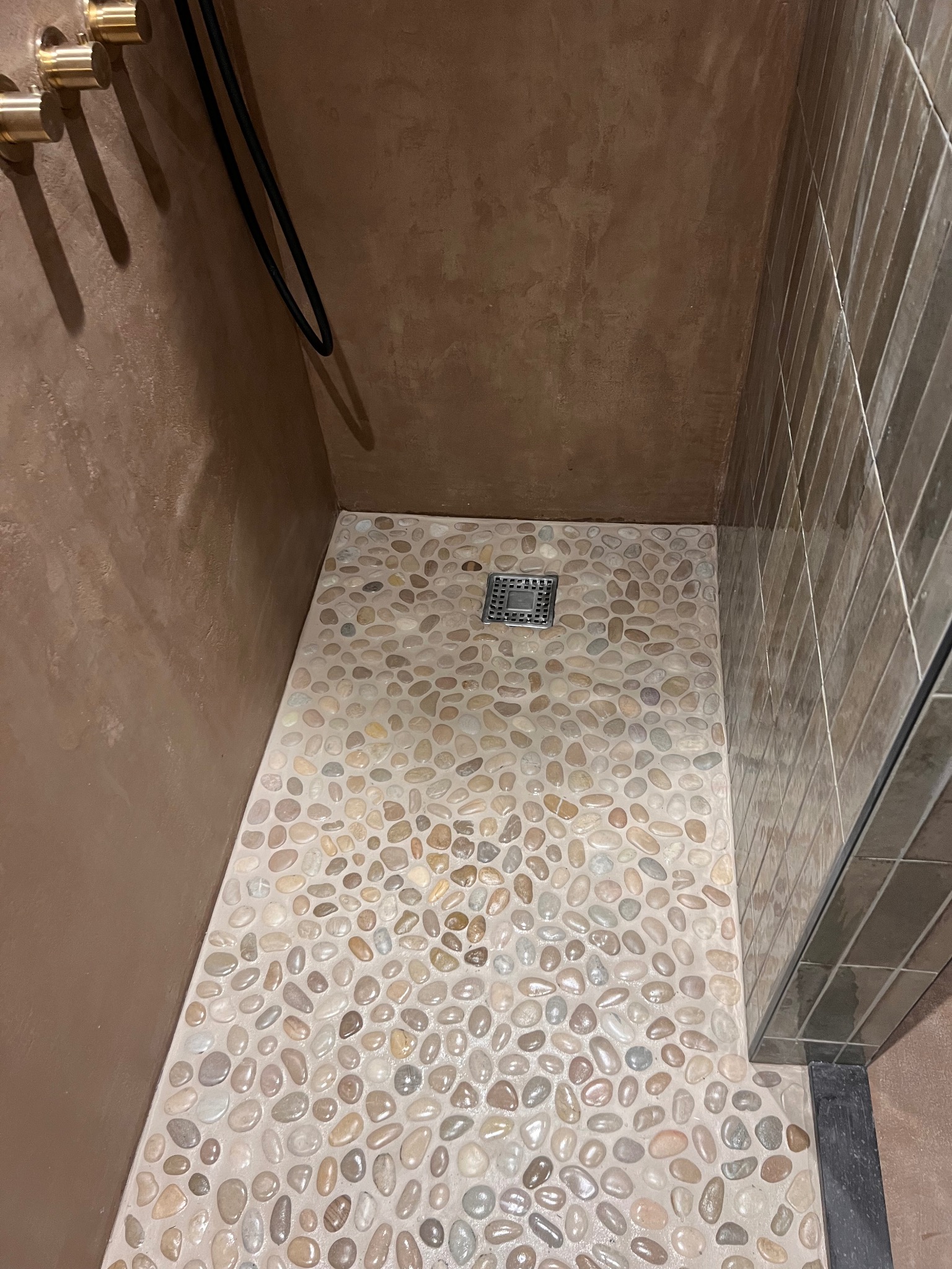 Kabina prysznicowa z podłogą wyłożoną drobnymi kamieniami w odcieniach beżu i brązu. Ściany wykończone w brązowym tynku i płytkach. Widoczny odpływ liniowy.