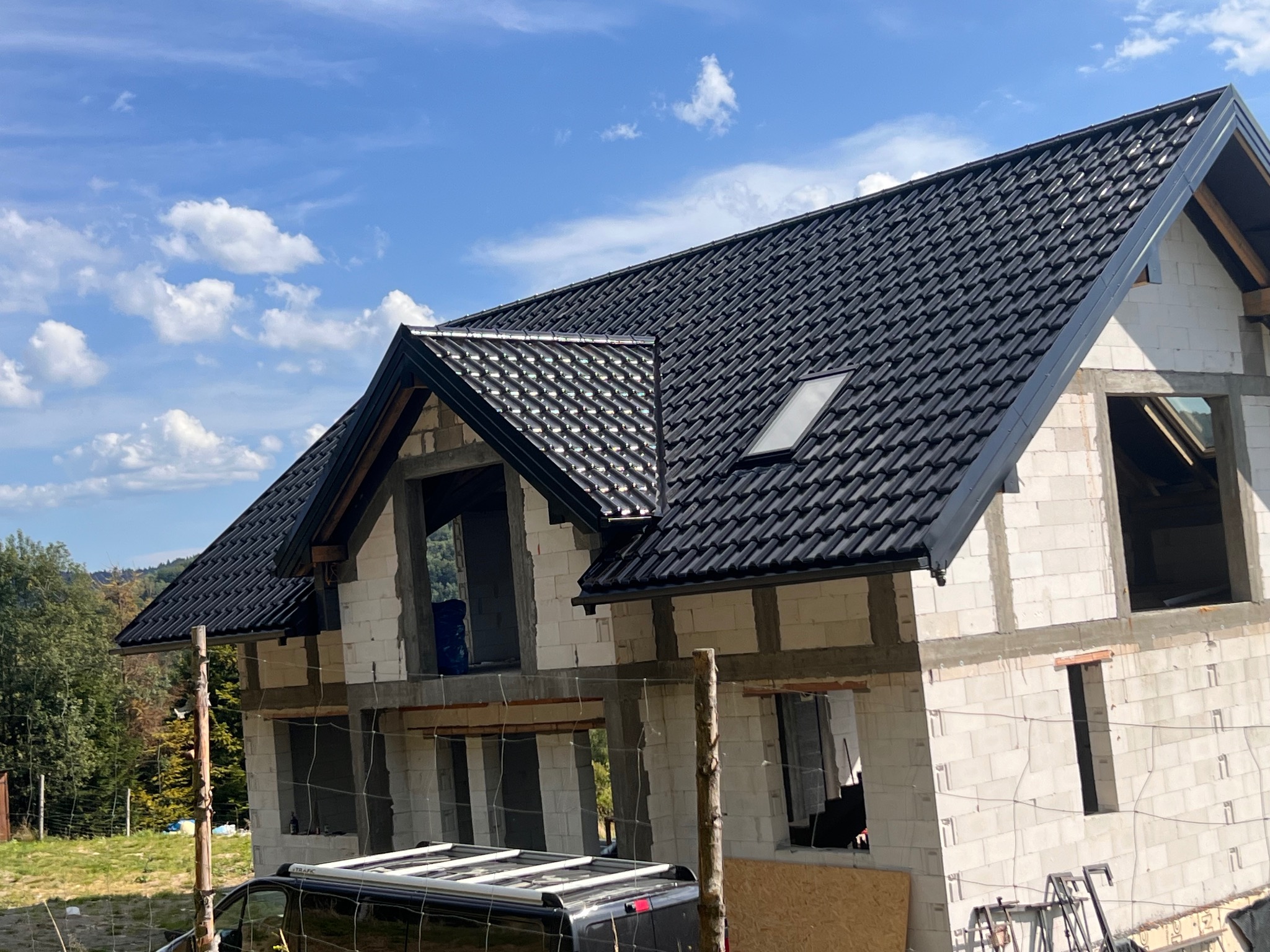 Dom w trakcie budowy z czarnym dachem z dachówki ceramicznej i białymi ścianami z betonu komórkowego, widok z dołu na tle błękitnego nieba.