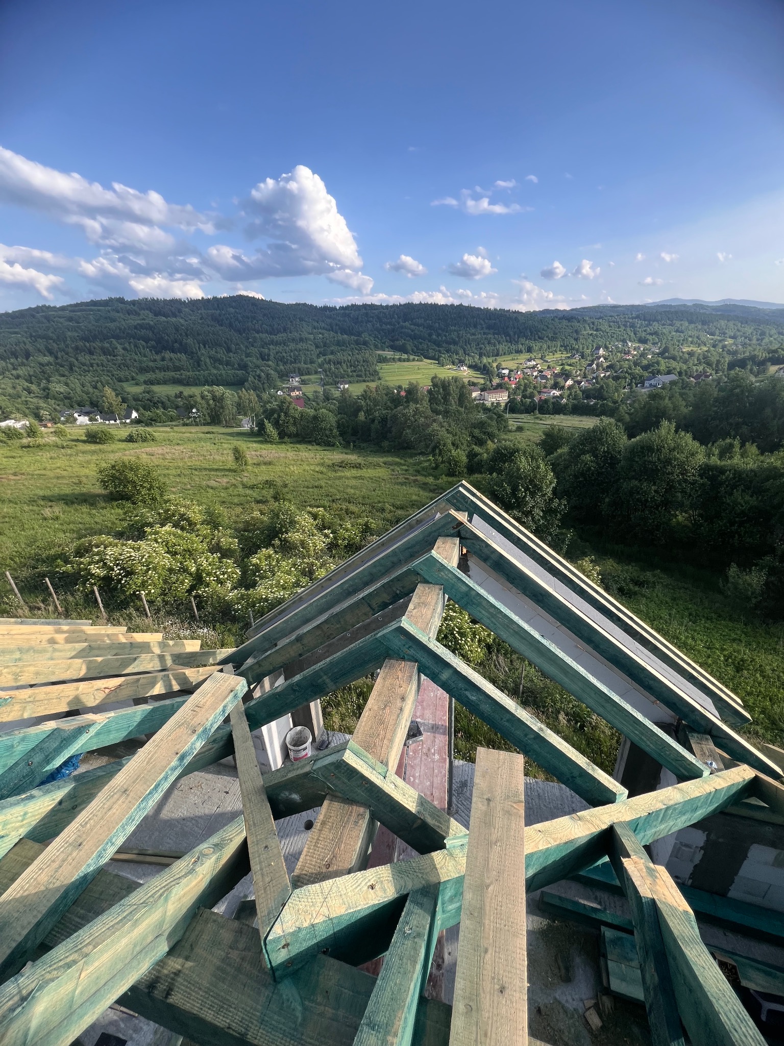 Konstrukcja dachu z drewnianych belek na tle malowniczego krajobrazu z zielonymi wzgórzami i błękitnym niebem z chmurami. Widok z góry na budowę.